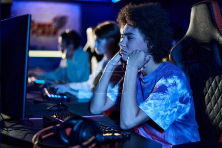 Téléchargez les photos : Triste femme afro-américaine regardant le moniteur d'ordinateur et gonflant les joues dans la pièce éclairée bleue - en image libre de droit