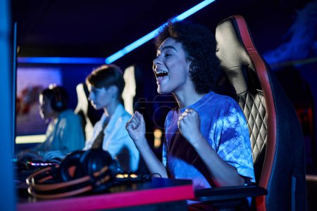 Téléchargez les photos : Excitée femme afro-américaine regardant l'écran d'ordinateur et acclamant dans la chambre éclairée bleue, gagnant - en image libre de droit
