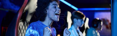 excited african american woman looking at computer monitor and cheering in blue lit room, banner