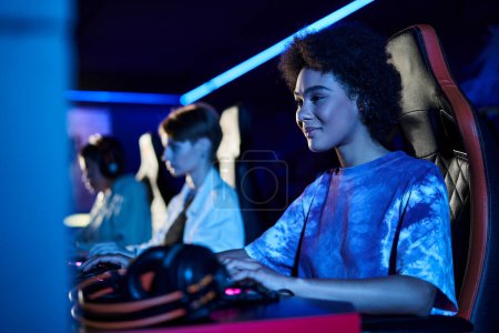 Téléchargez les photos : Heureux afro-américaine femme regardant moniteur dans la chambre éclairée bleue, jouer jeu d'ordinateur - en image libre de droit