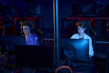 diverse female friends in headphones looking at monitors while playing multiplayer computer game