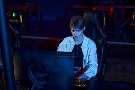 mujer enfocada mirando el monitor mientras juega juego de ordenador multijugador, jugador en la habitación iluminada azul