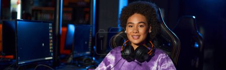 happy african american female gamer with headphones sitting on comfortable gaming chair, banner