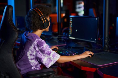 Foto de Jugador americano africano rizado en auriculares jugando juego de ordenador mientras mira la pantalla, cybersport - Imagen libre de derechos