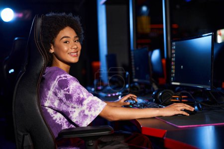 femme afro-américaine joyeuse assise sur une chaise de jeu et regardant la caméra, le cybersport