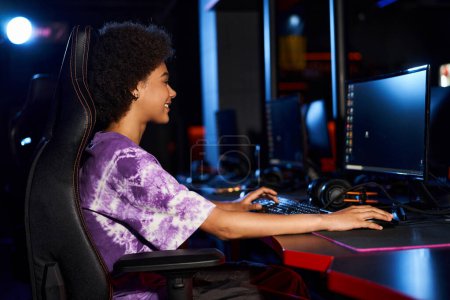 Foto de Vista lateral de la mujer afroamericana feliz sentado en silla de juego y mirando el monitor, cybersport - Imagen libre de derechos