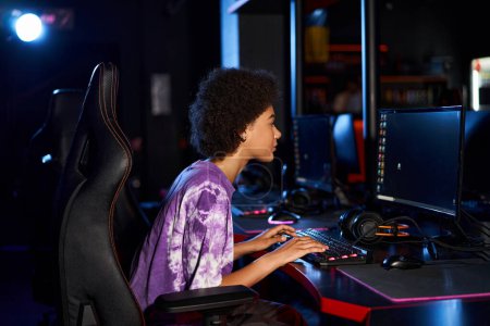Foto de Jugador americano africano rizado en auriculares jugando juego de ordenador mientras mira la pantalla, cybersport - Imagen libre de derechos