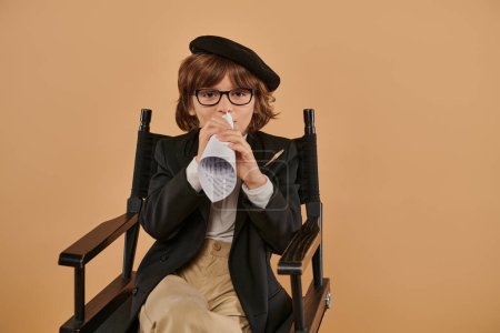 Stilvoller Junge mit Baskenmütze und Brille sitzt im Regiestuhl und spricht in einem Stück Papier