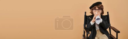 Photo for Stylish boy in beret and glasses sits in director chair, speaking in piece of rolled paper, banner - Royalty Free Image