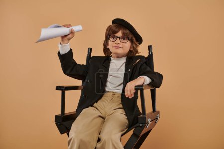 Junge, stilvoll gekleidet, sitzt auf dem Regiestuhl mit gerolltem Papier und einem Lächeln im Gesicht