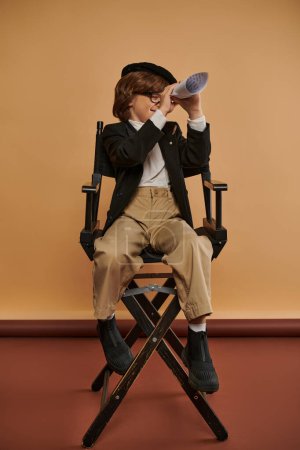mignon garçon en vêtements élégants se trouve sur la chaise du directeur et regarder à travers le trou dans le papier laminé