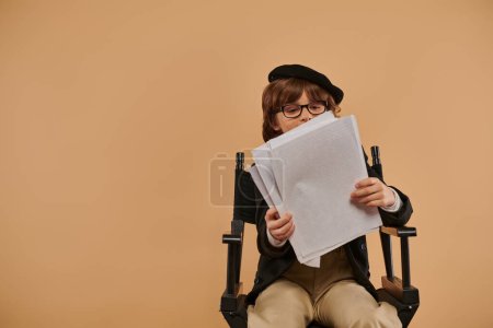 busy boy in glasses sits in director chair, looking at papers and absorbed in his reading