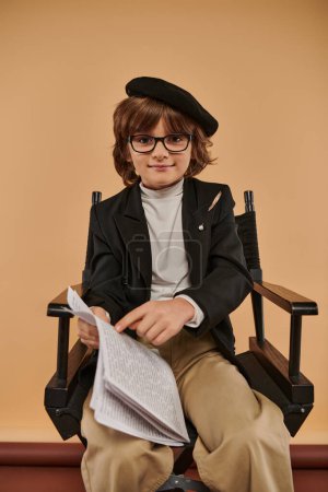 niño en boina y gafas se sienta en silla de director, señalando con el dedo en el papel, profesión y niño