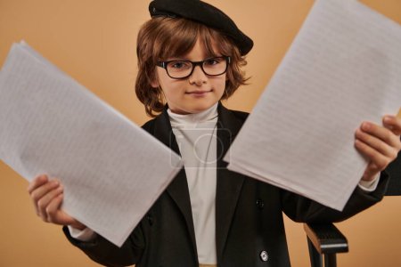 boy dressed in stylish clothing and wearing glasses stands confidently with papers in hands