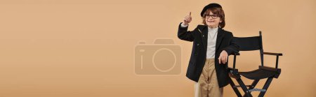 Photo for Joyful boy standing near director chair, with a smile on face showing idea sign,  profession banner - Royalty Free Image