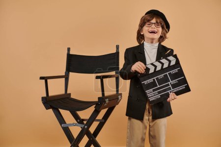 excited filmmaker boy in trendy clothing happily poses with clapperboard in hand against beige wall