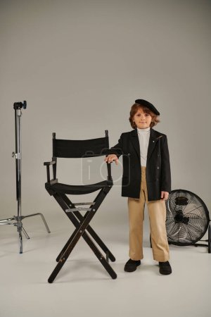 stylish boy in beret and smart casual attire stands confidently near director chair on grey backdrop