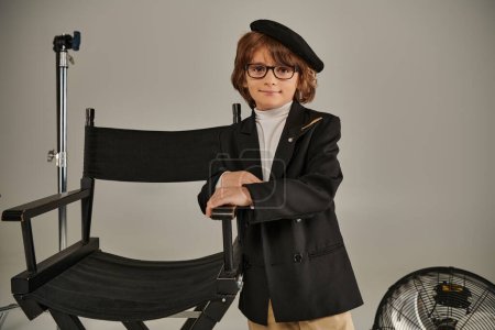 Netter Junge in Baskenmütze und stylischer Kleidung steht selbstbewusst neben dem Regiestuhl auf grauem Hintergrund