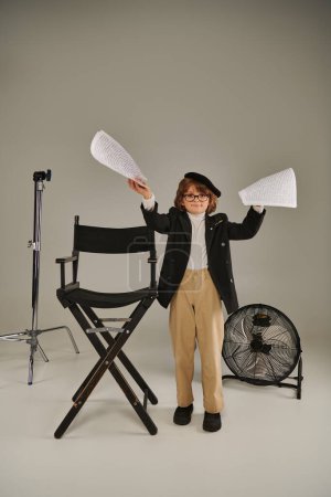 stylischer Junge mit Brille und Baskenmütze mit Drehbuch auf Papier auf grau, Kind als Regisseur