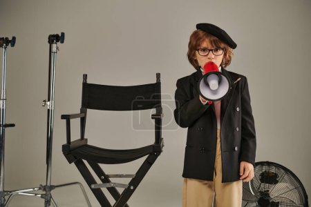 Junge als Filmemacher steht selbstbewusst neben Regiestuhl und spricht in Megafon, grauem Hintergrund