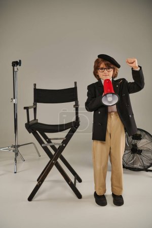 A young activist in beret stands with megaphone and director chair on grey, boy as filmmaker