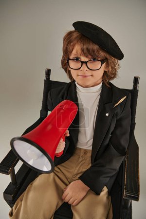 cute filmmaker boy in beret and glasses sitting on director chair and holding red megaphone