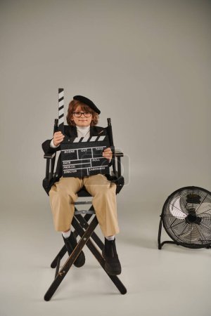 boy in beret and glasses holding clapper board as sitting on director chair, film making concept