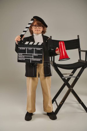 chico en boina y gafas celebración clapper tablero y rojo megáfono y de pie cerca de director silla