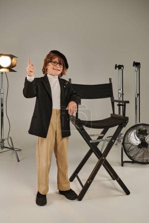 feliz chico con estilo en boina y gafas de pie delante de la silla del director, softbot en el fondo