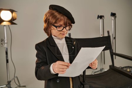 Stilvolles Kind mit Brille und Baskenmütze liest Drehbuch auf Papier, Junge als Regisseur des Films