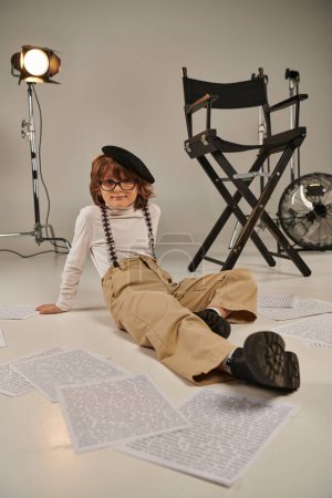 garçon dans les bretelles et béret assis sur le sol entouré par le scénario, garçon en tant que réalisateur de cinéaste