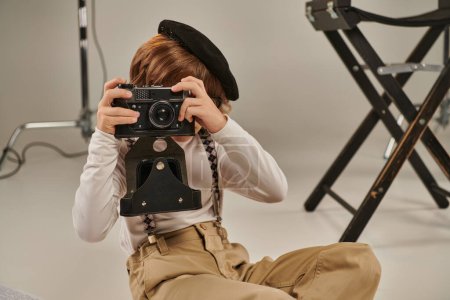 niño captura el momento en la cámara retro y sentado en el suelo cerca de la silla de director, joven fotógrafo