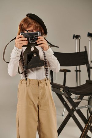 Junge in Baskenmütze fängt den Moment mit Retrokamera in der Nähe des Regiestuhls ein, junger Fotograf im Studio