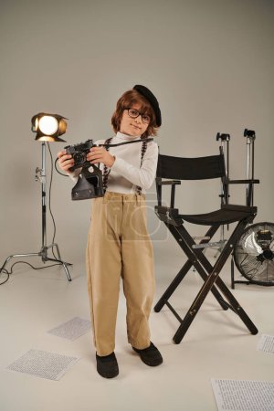 Junge mit Baskenmütze und Brille fängt den Moment mit Retrokamera in der Nähe des Regiestuhls ein, junger Fotograf