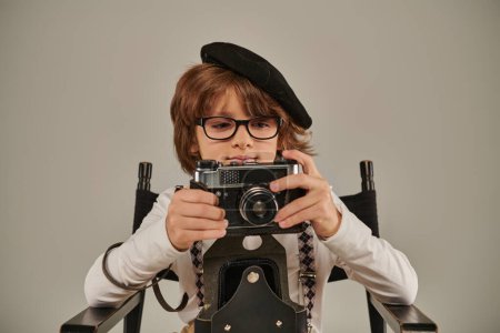Kind in Baskenmütze und Brille mit Retro-Kamera auf dem Regiestuhl sitzend, junger Fotograf