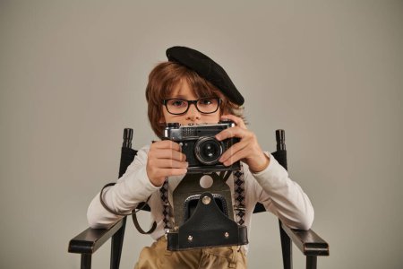 niño en boina y gafas sosteniendo la cámara vintage mientras está sentado en la silla del director, joven fotógrafo