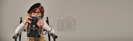 niño en boina tomando fotos en la cámara vintage mientras está sentado en la silla del director, cartel del fotógrafo
