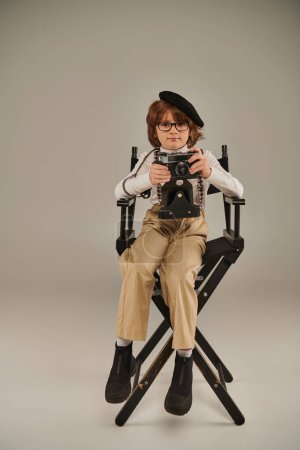young photographer in beret and glasses holding retro camera while sitting on director chair