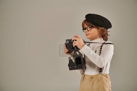 niedlicher Junge, junger Fotograf in Baskenmütze und Hosenträgern, der im Studio mit der Retro-Kamera fotografiert
