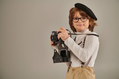 Junge, junge Fotografin in Baskenmütze und Hosenträgern beim Fotografieren mit Retro-Kamera im Studio