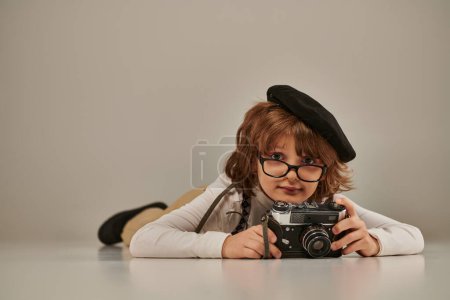 happy young photographer in beret and suspenders taking photo on retro camera and lying on floor