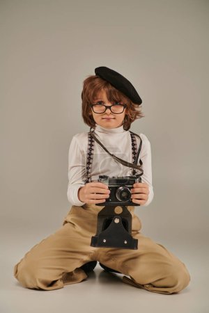 young photographer in beret and glasses holding camera and sitting on floor, boy in suspenders