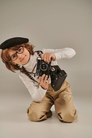 junger Fotograf in Baskenmütze mit Retro-Kamera auf dem Boden sitzend, neugieriger Junge in Hosenträgern