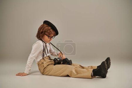 young photographer in beret and glasses holding camera and sitting on floor, cute boy in suspenders