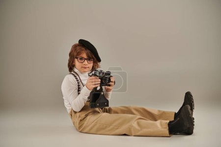 joven fotógrafo en boina y gafas sosteniendo la cámara y sentado en el suelo, lindo niño en tirantes