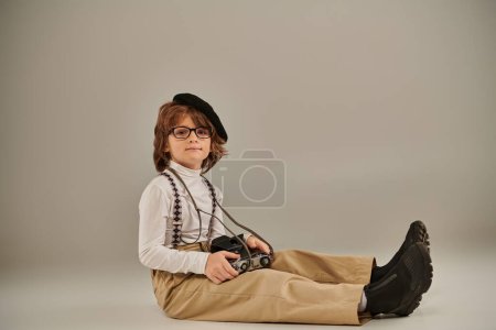 junger Fotograf in Baskenmütze und Brille mit Retro-Kamera auf dem Boden sitzend, Kind in Hosenträgern