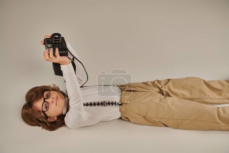 niño feliz captura un momento mientras está acostado en el suelo, niño en boina y gafas con cámara retro