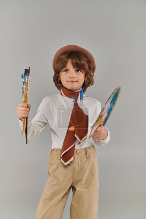 young painter holding paint brush and palette, boy in beret and scarf standing on grey backdrop