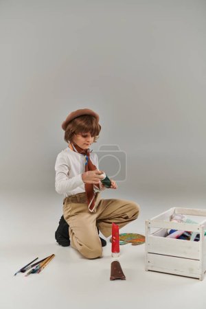 boy kneels on the floor, surrounded by paints in tubes and a wooden tool box, young painter in beret