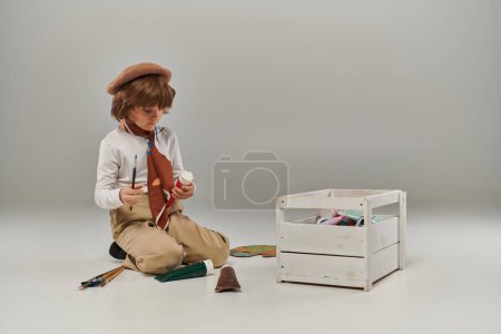 boy kneels on the floor surrounded by paints in tubes and a wooden tool box, young artist in beret